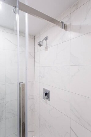Eine begehbare, gläserne Dusche mit Handbrause, weißen Marmorfliesen und sichtbarer Bedienung.