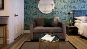 A room with a sofa, a round coffee table with a book, a bed with white bedding, a nightstand with a lamp, and a wall with a palm leaf pattern. - 6768 - 203 · Golden Oasis mini Studio with full bath  by Denstays (Montréal)