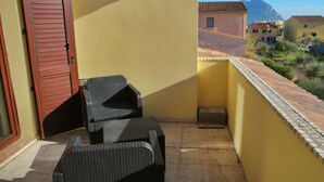 Un balcón con mobiliario de mimbre, una puerta de madera cerrada y vistas a edificios y montañas.