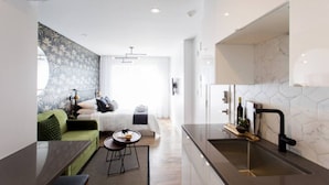 Una habitación de hotel moderna con una cama, un sofá verde, una mesita y una zona de cocina con un lavabo y encimera.