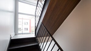 Una escalera con barandilla de madera y balaustradas de metal, que lleva a un ventanal con vistas a edificios.