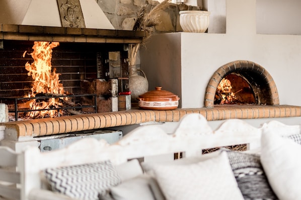 A brick fireplace with a fire, a stone oven, and a white cushioned seating area.