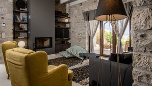A cozy living room with a stone wall, a fireplace, a large window with curtains, and a modern sofa. - Casa Della Vita (Hersonissos)