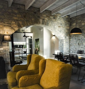 A cozy interior with a stone archway leading to a kitchen and dining area, featuring a mustard-colored armchair and a hanging pendant light.