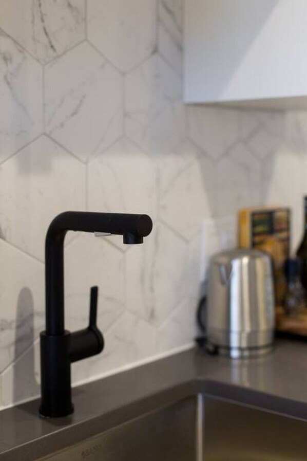 A kitchen with a modern faucet, a stainless steel kettle, and a marble backsplash.