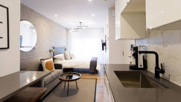 Une chambre d’hôtel moderne dotée d’une kitchenette, d’un lit avec une tête de lit, d’une table basse ronde et d’une grande fenêtre.