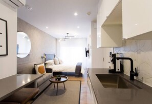 A modern hotel room with a kitchenette, a bed with a headboard, a round coffee table, and a large window.