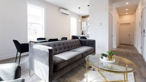 Een moderne woonkamer met een grijze bank, zwarte stoelen, een glazen salontafel en een eethoek met een witte tafel en stoelen.