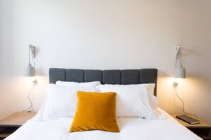 A neatly made bed with a grey upholstered headboard, white and mustard pillows, and two bedside lamps on nightstands.
