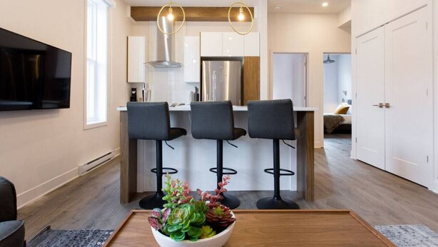 A modern kitchen with a central island, bar stools, pendant lighting, and a flat-screen TV mounted on the wall. - Brand New Light filled Mile End Flat by Denstays (Montréal)