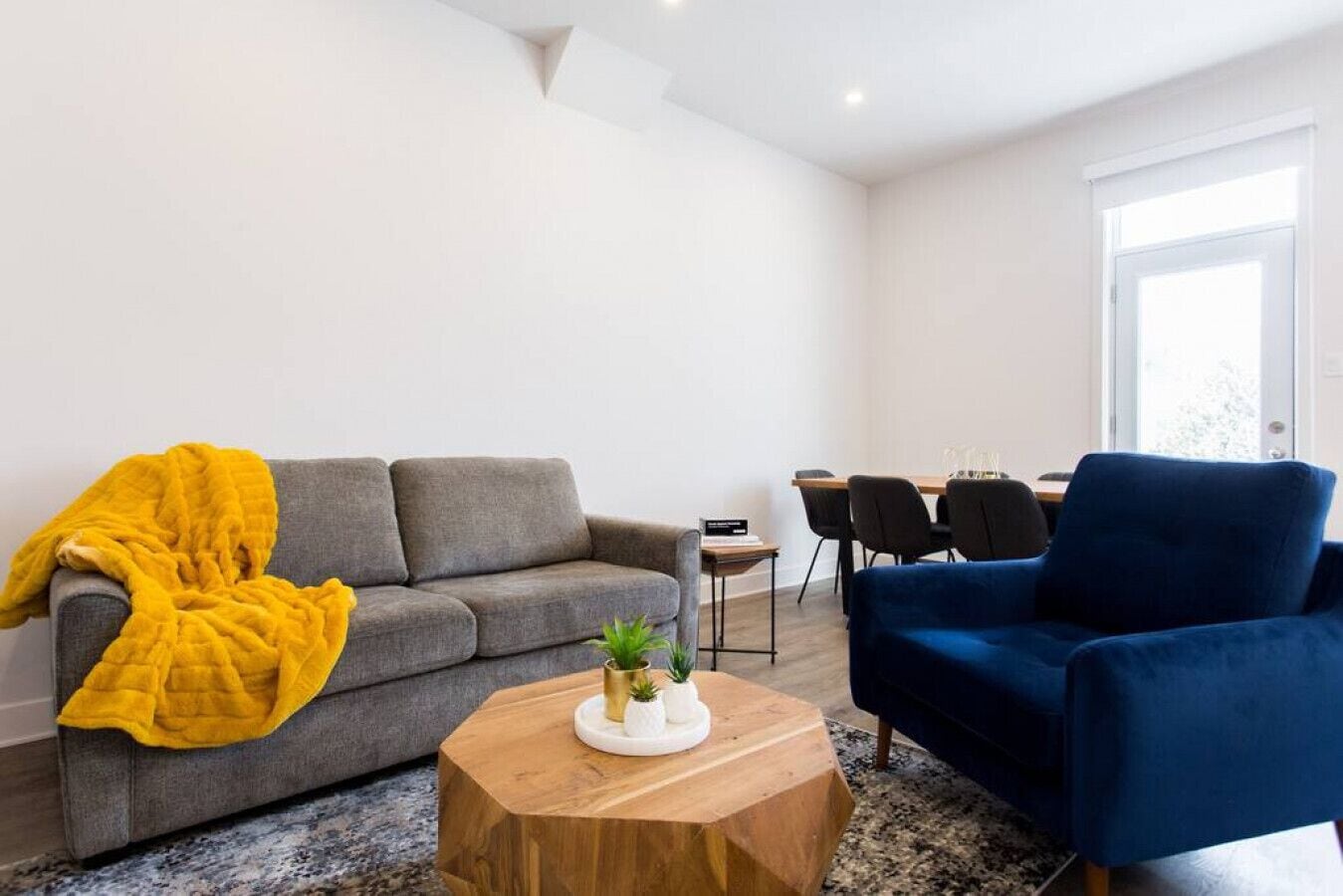 Un salon moderne avec un canapé gris, un fauteuil bleu, une table basse en bois et, au fond, un coin repas.