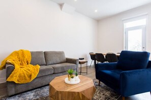 Un salon moderne avec un canapé gris, un fauteuil bleu, une table basse en bois et, au fond, un coin repas.