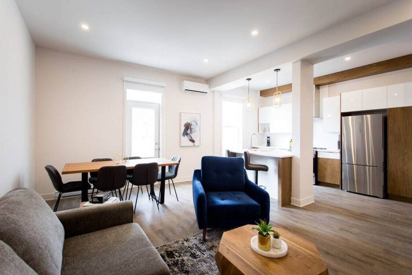 A modern living room with a sofa, a dining table with chairs, a blue armchair, and a kitchen area with a refrigerator and a sink.