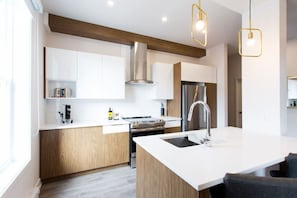 Une cuisine moderne avec des meubles blancs, une cuisinière en acier inoxydable, un îlot central doté d’un évier et des luminaires suspendus.