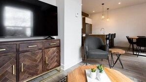 A modern living room with a flat-screen TV, a herringbone-patterned wooden cabinet, a grey armchair, a wooden coffee table with plants, and a dining area with a bar stool and chairs. - Light Filled 2 Bdr Unit in Mile End by Denstays (Montréal)