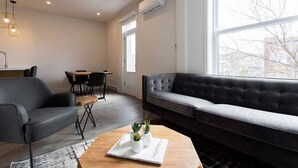 A modern living room with a grey sofa, a wooden coffee table with plants, a dining area with a table and chairs, and large windows. - Light Filled 2 Bdr Unit in Mile End by Denstays (Montréal)
