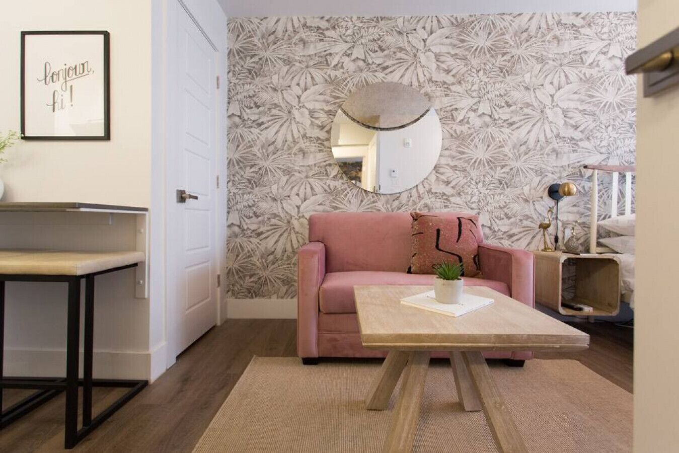 Ein Zimmer mit einem rosa Sofa, einem runden Spiegel, einem kleinen Tisch mit einer Pflanze und einer Wand mit floralem Muster.