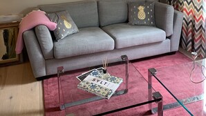 Un soggiorno con un divano grigio, una coperta rosa, un tavolino da caffè in vetro e riviste, e un quadro che raffigura un cervo appeso al muro.