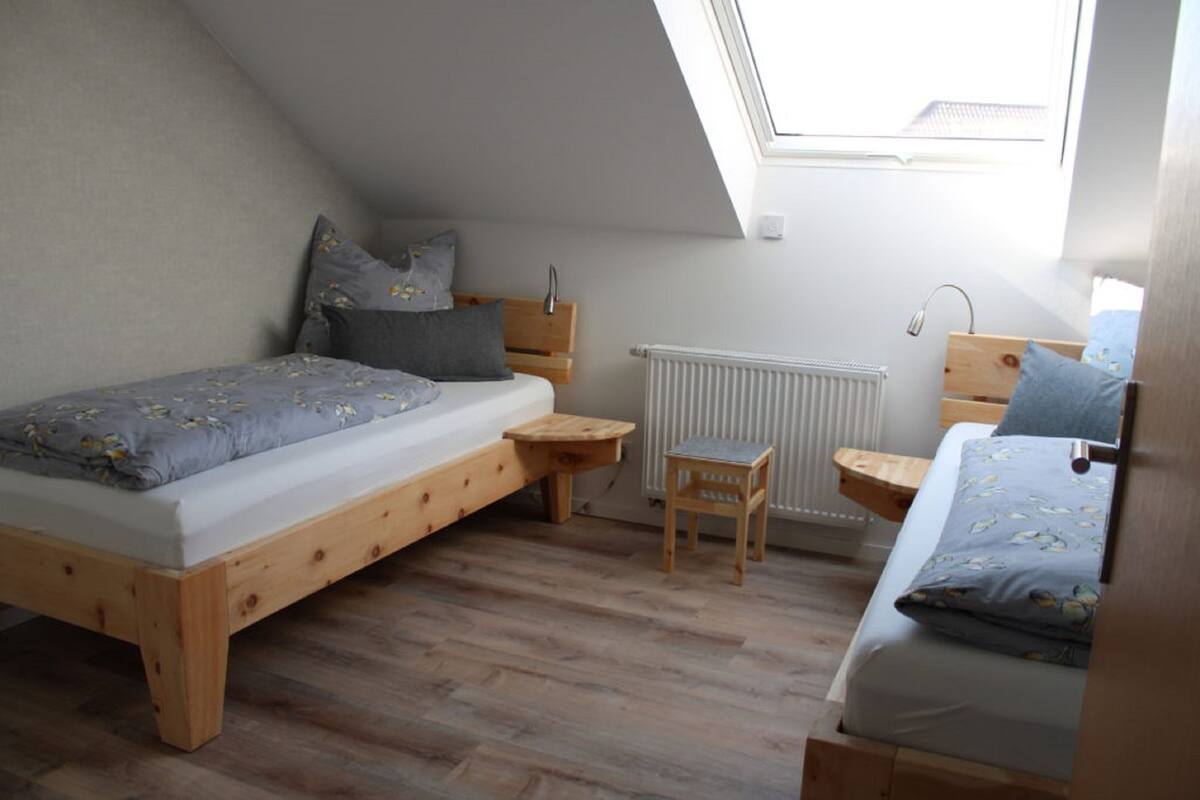 Quarto com teto inclinado, um claraboia, cama de madeira, mesa de madeira pequena, cadeira e radiador.