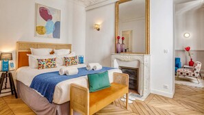 A hotel room with a bed, a bench, a fireplace, a mirror, and a painting on the wall.