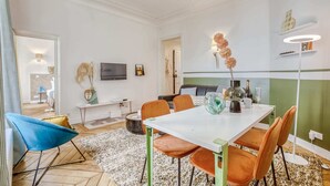 Un salon moderne avec une table à manger blanche, des chaises orange, un fauteuil bleu et un téléviseur fixé au mur.