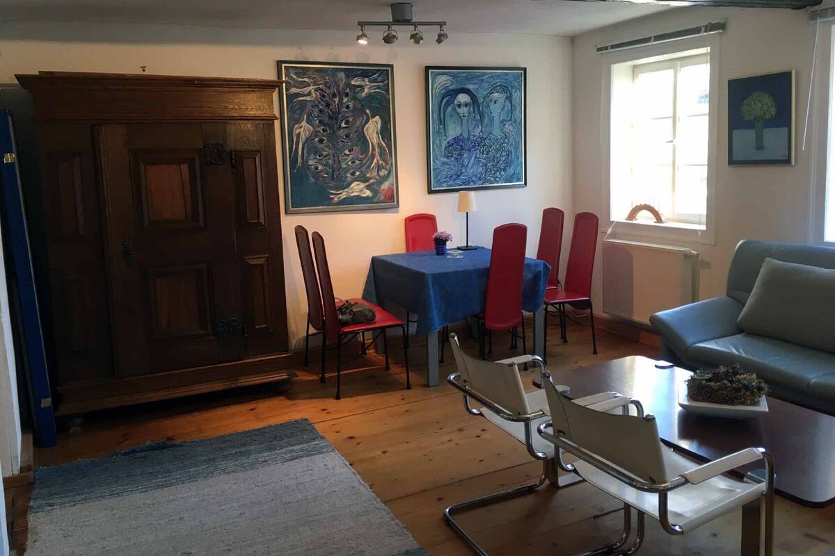 Un coin repas avec une table et des chaises, deux tableaux accrochés au mur, une fenêtre avec des rideaux et un canapé placé dans un coin.