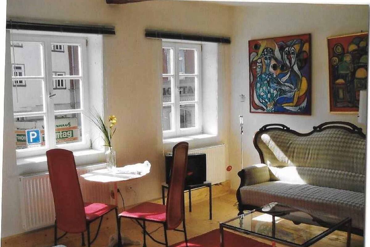 Une pièce comprenant un canapé, deux fauteuils, une petite table et un radiateur. Il y a deux fenêtres donnant sur la rue, et un tableau accroché au mur.
