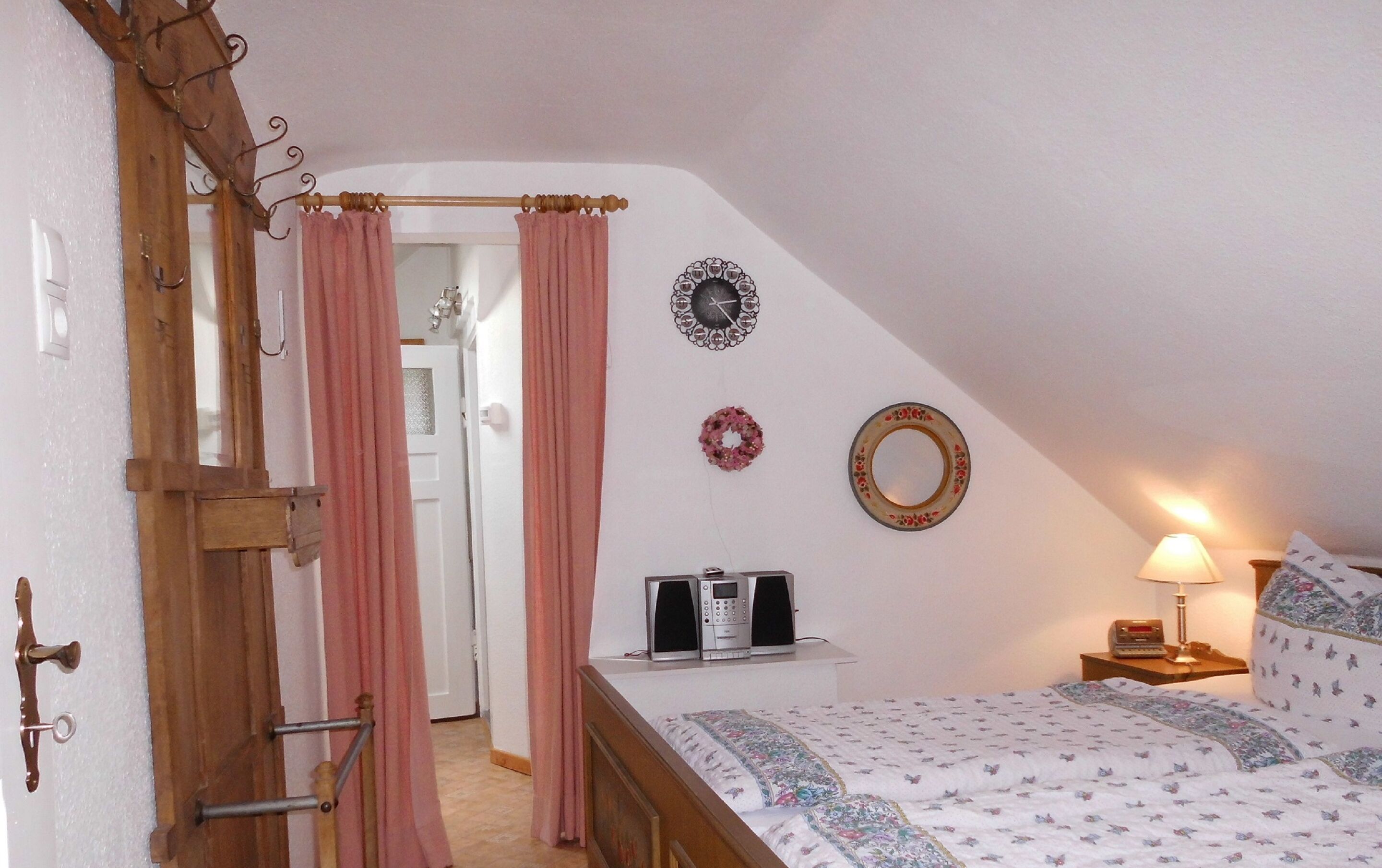 A bedroom with a bed, a bedside table with a lamp, a wall clock, and a door with curtains.