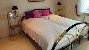 A neatly made bed with a white comforter and multiple pillows, flanked by two gold-framed bedside tables with lamps, in a room with a simple, clean design.