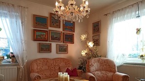 A cozy living room with a large octagonal table, two patterned armchairs, a chandelier, and several framed artworks on the walls.