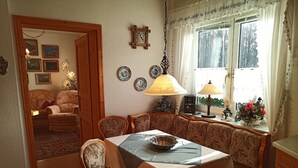 A dining area with a table set for four, surrounded by patterned chairs. There are two wall-mounted lamps, a clock, and a window with curtains.