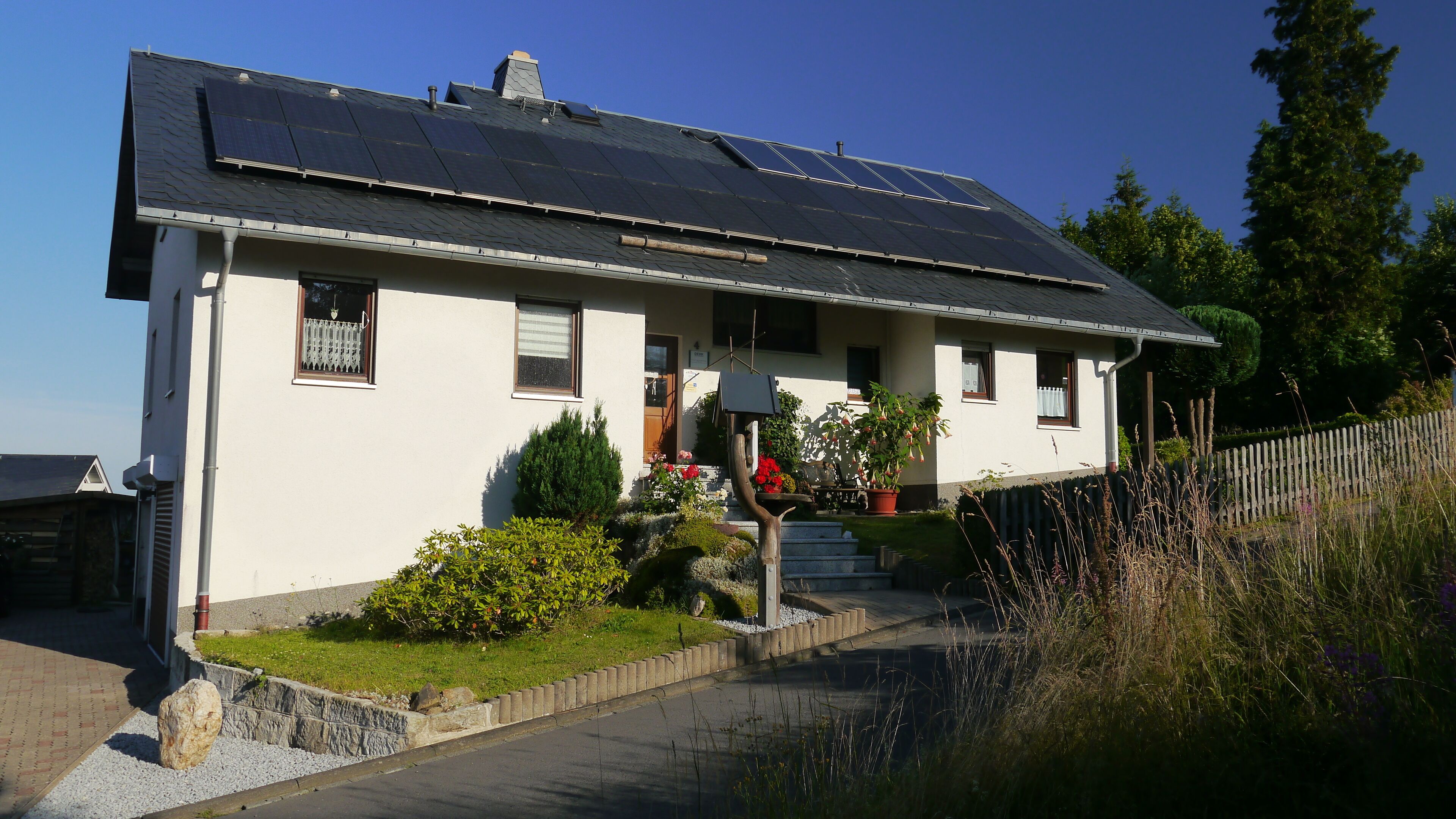Una casa con paneles solares en el techo, un pequeño jardín con flores y un camino de entrada pavimentado.