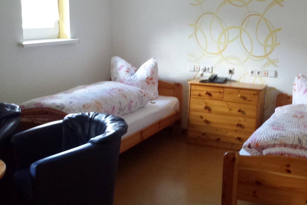 Une petite pièce bien éclairée, avec deux lits simples, une commode en bois et une chaise.