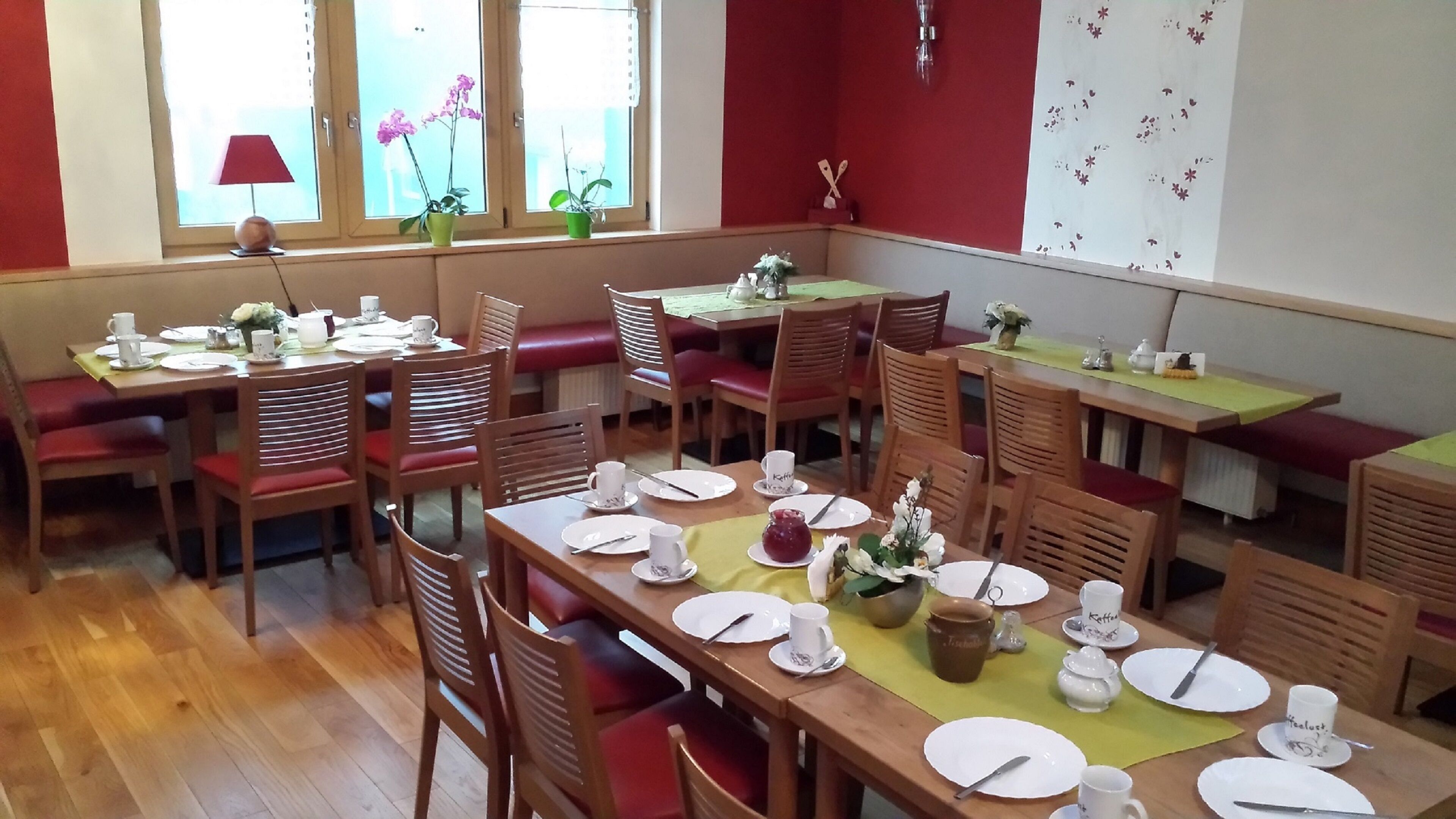 Un espace repas avec plusieurs tables et chaises, dont certaines sont dotées de coussins rouges. On trouve des plantes en pot sur le rebord de la fenêtre et un mur orné d’un motif floral.