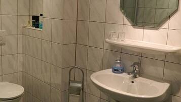Een badkamer met toilet, wastafel en douche. Er is een plank met toiletartikelen en een spiegel boven de wastafel.