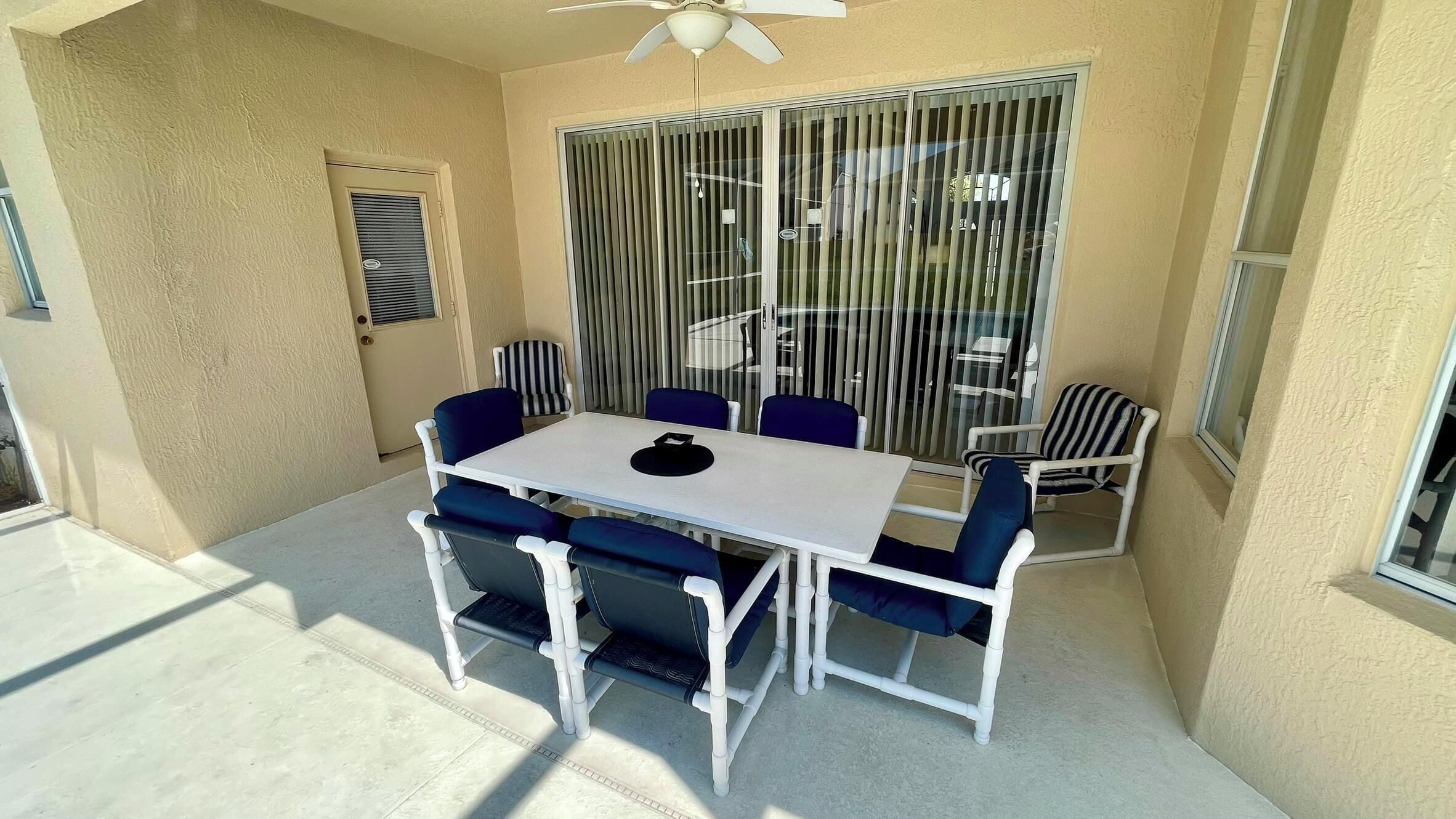 A patio with a white table and blue chairs, a ceiling fan, and a sliding glass door with vertical blinds.