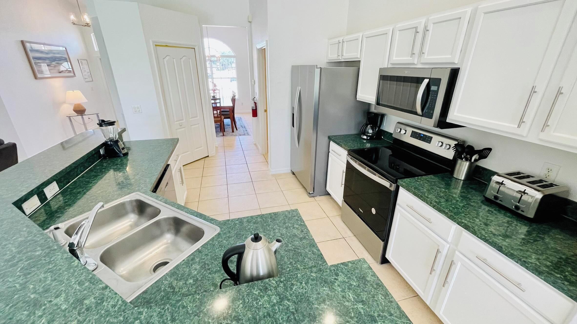 A kitchen with white cabinets, a stainless steel sink, a kettle, a microwave, a refrigerator, and a toaster.