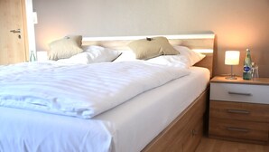 A neatly made bed with white linens and two beige pillows, a wooden nightstand with a lamp and a bottle, and a wooden drawer unit.