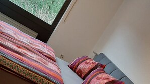 Una cama con un edredón colorido y dos almohadas, una ventana con vistas a follaje, y un armario de madera.