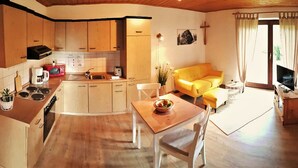 Eine kompakte Küche mit Holzausstattung, einem Esstisch mit Obstschale und einem gelben Sofa in einem hellen Wohnbereich mit Fernseher und Kruzifix an der Wand.