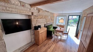 A cozy room with a stone wall, wooden floor, a flat-screen TV, a wooden cabinet, a green sofa, and a dining table with chairs. - Residenz Nr.6 (Zapfendorf)