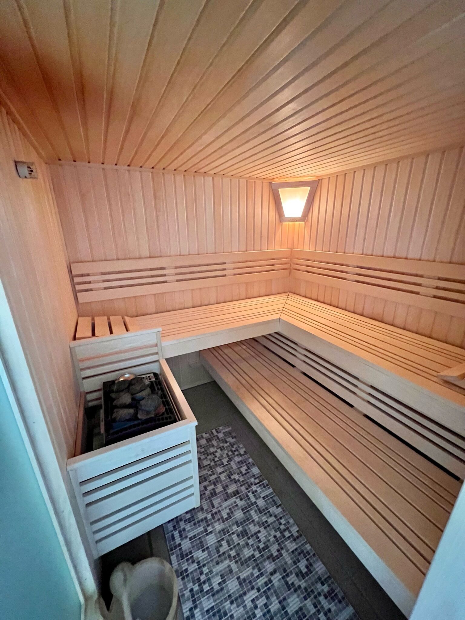 A sauna with wooden benches and a small window. There is a storage area with a metal rack and a mat on the floor.