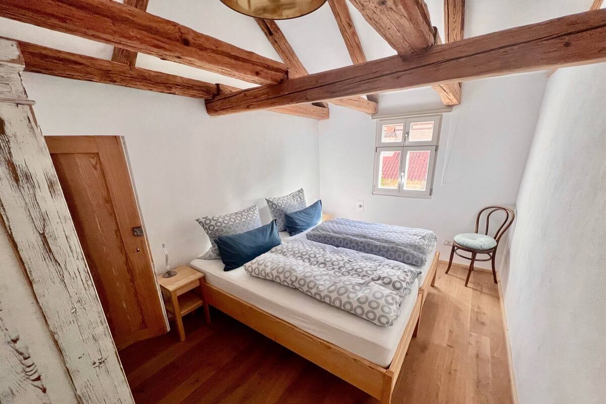 A bedroom with a wooden bed, a small bedside table, a chair, and a window with a view of the outside.