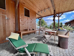 A wooden cabin with a covered patio area featuring two green lounge chairs, a wooden table, and a stone fireplace. - Residenz Nr.6 (Zapfendorf)