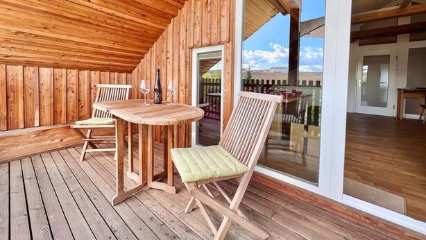 A wooden deck with a round table and chairs, a glass door leading outside, and a view of a building in the background. - Residenz Nr.6 (Zapfendorf)