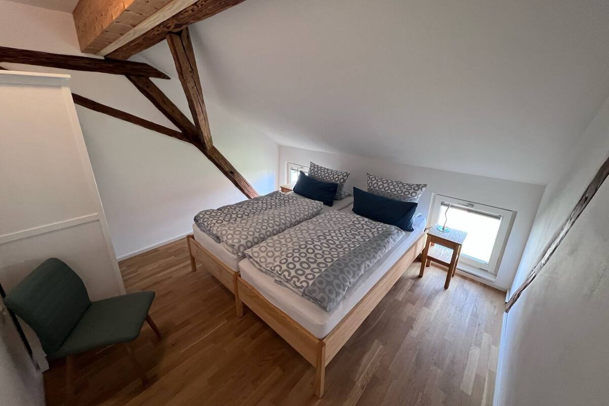 A loft bedroom with a wooden beam ceiling, a bed with patterned bedding, a chair, and a small table.