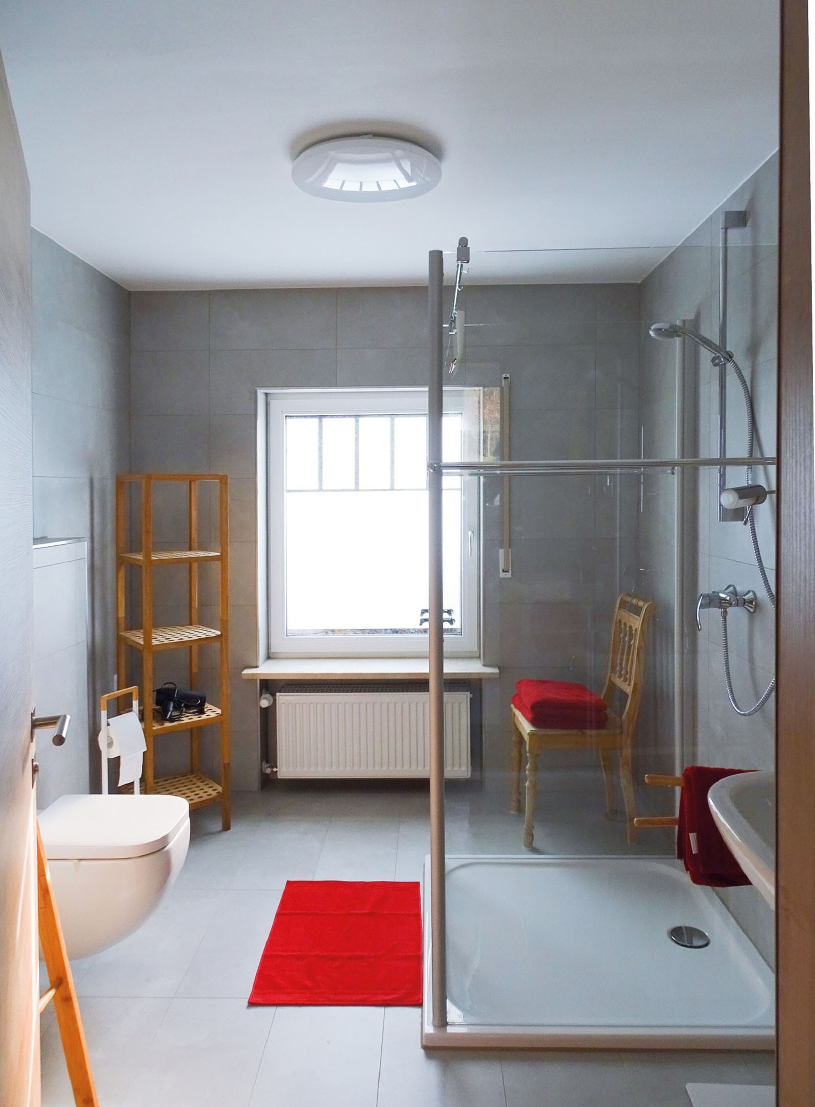 Un baño moderno con cabina de ducha de vidrio, una silla de madera, un tapete rojo y un ventanal que proporciona luz natural.