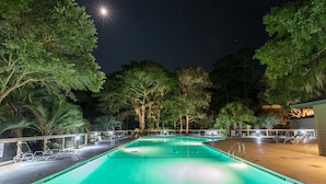 A nighttime swimming pool with clear water, surrounded by trees and lounge chairs.