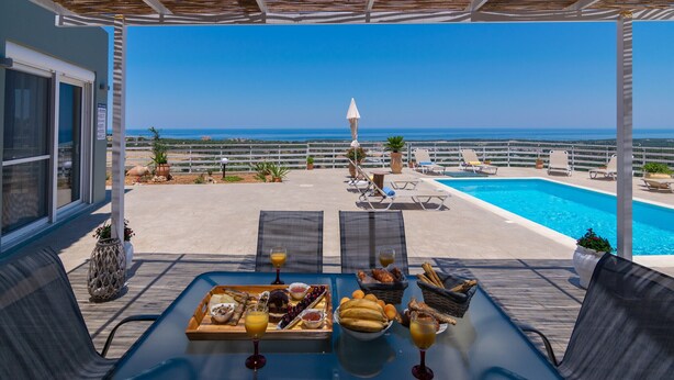 A terrace with a pool, dining table with breakfast, lounge chairs, and a view of the sea.