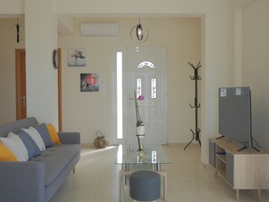 A living room with a sofa, a glass coffee table, a TV stand, and a coat rack. - Haravgi villa 3 ,near resort,private pool (Agia Triada)